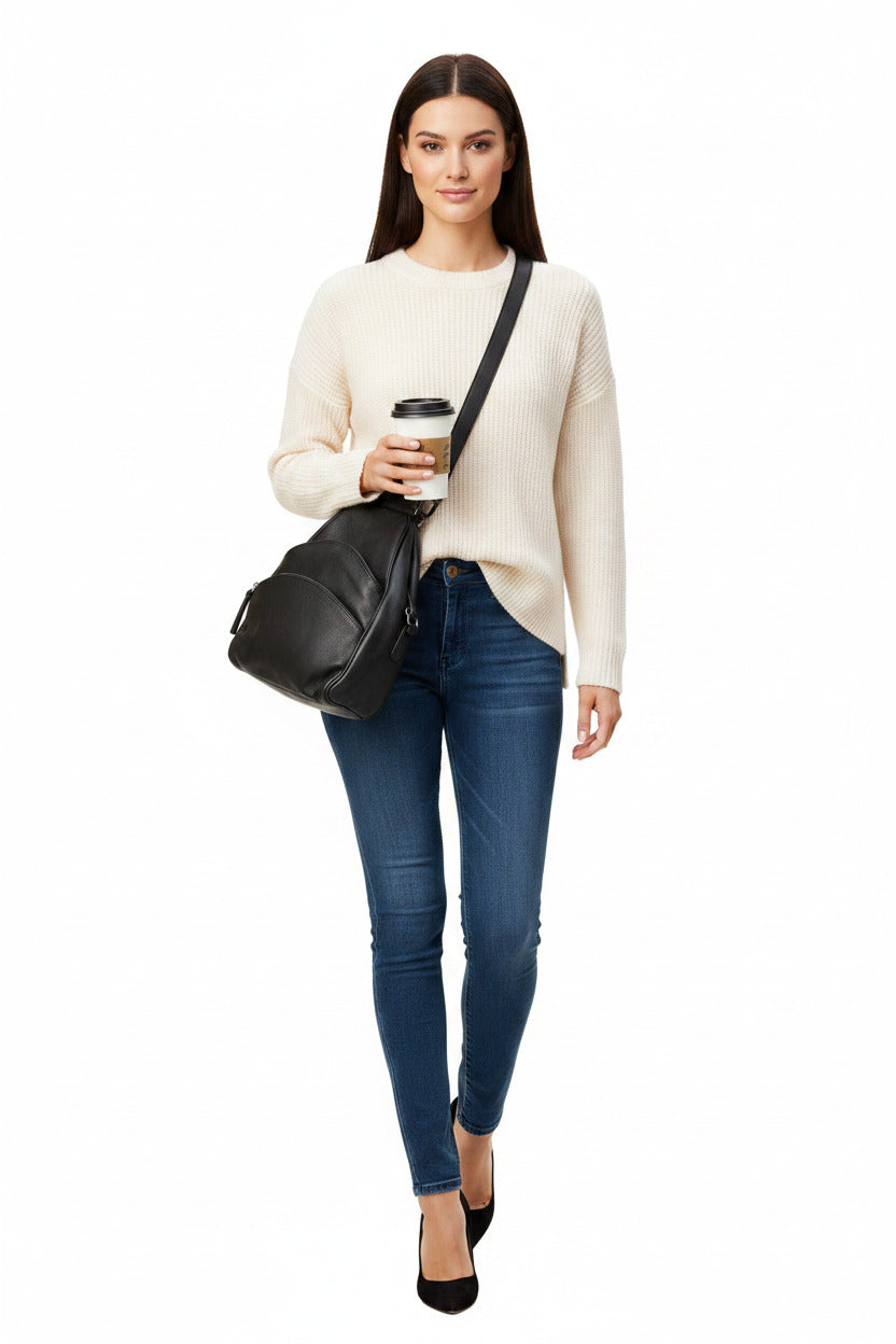 A woman wearing sunglasses, a cream knit sweater, and blue jeans carries a black leather crossbody bag and holds a takeaway coffee cup against a plain white background.