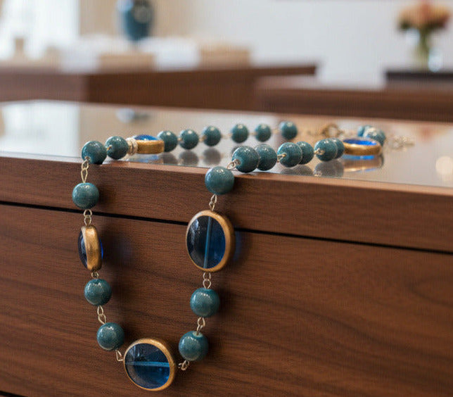 A beaded necklace featuring marbled teal blue beads and two large oval blue glass inserts with gold-toned metal trim, shown against a plain white background.