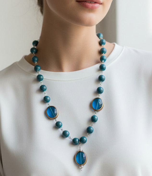 A beaded necklace featuring marbled teal blue beads and two large oval blue glass inserts with gold-toned metal trim, shown against a plain white background.