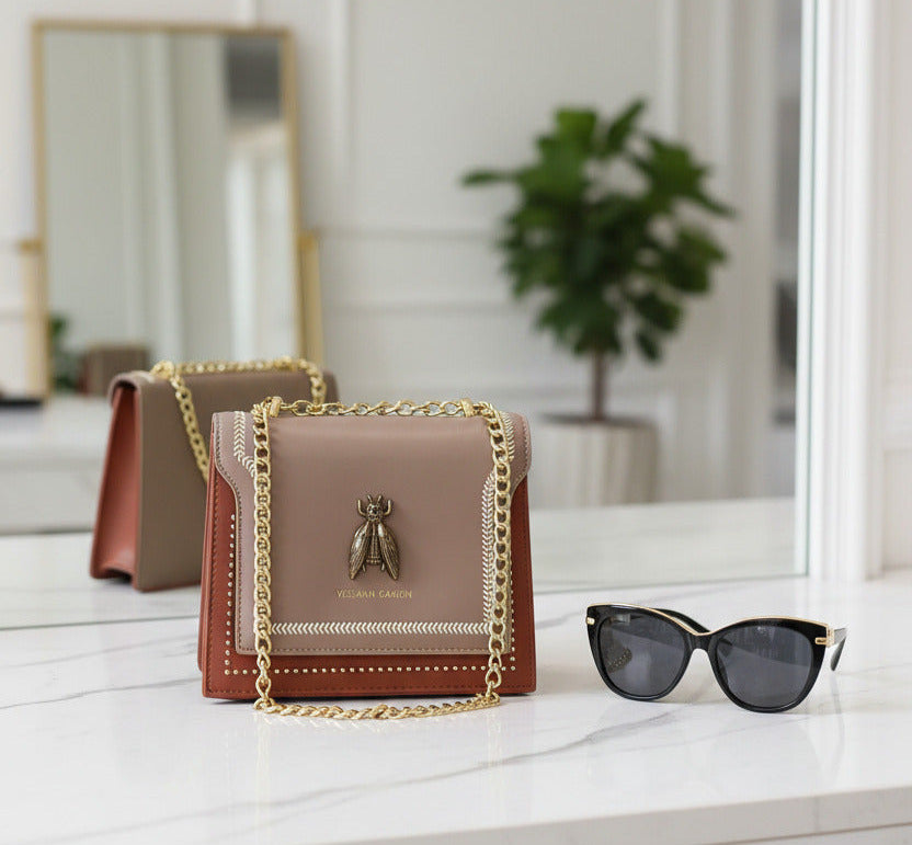 A small Vessana Gahon shoulder bag in taupe and brown leather with gold-tone chain strap and a bee hardware accent on the front flap, shown against a plain white background.