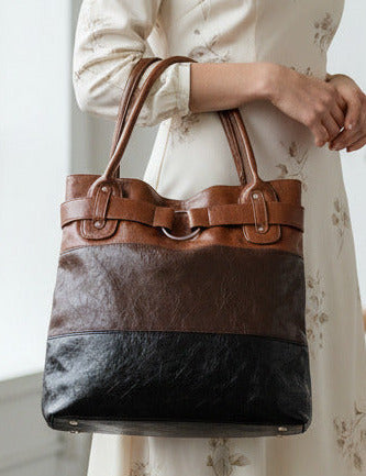 A woman stands in a bright room holding a large color-block tote bag. The bag has a black lower section, a dark brown middle section, and a tan upper section with double shoulder straps.