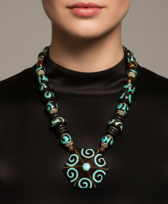 A beaded necklace featuring a large hexagonal pendant with turquoise and brown spiral patterns, flanked by dark brown and turquoise beads with metallic spacer details, shown against a plain white background.