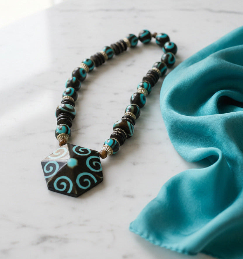 A beaded necklace featuring a large hexagonal pendant with turquoise and brown spiral patterns, flanked by dark brown and turquoise beads with metallic spacer details, shown against a plain white background.
