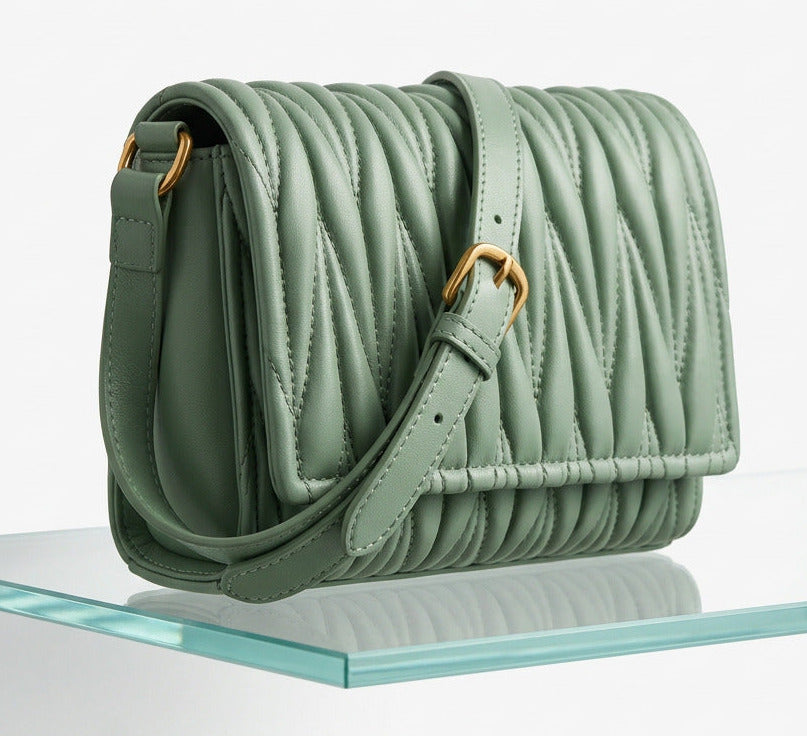 A sage green shoulder bag on a glass shelf displayed against white background.