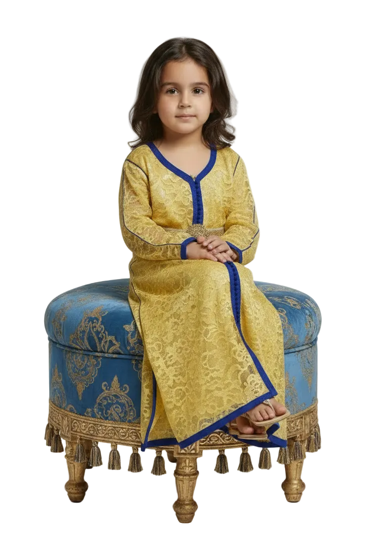 A young girl modeling a long-sleeved golden-yellow kaftan dress with blue trim, a matching belt, and a decorative gold medallion at the waist, set against a plain white studio background.