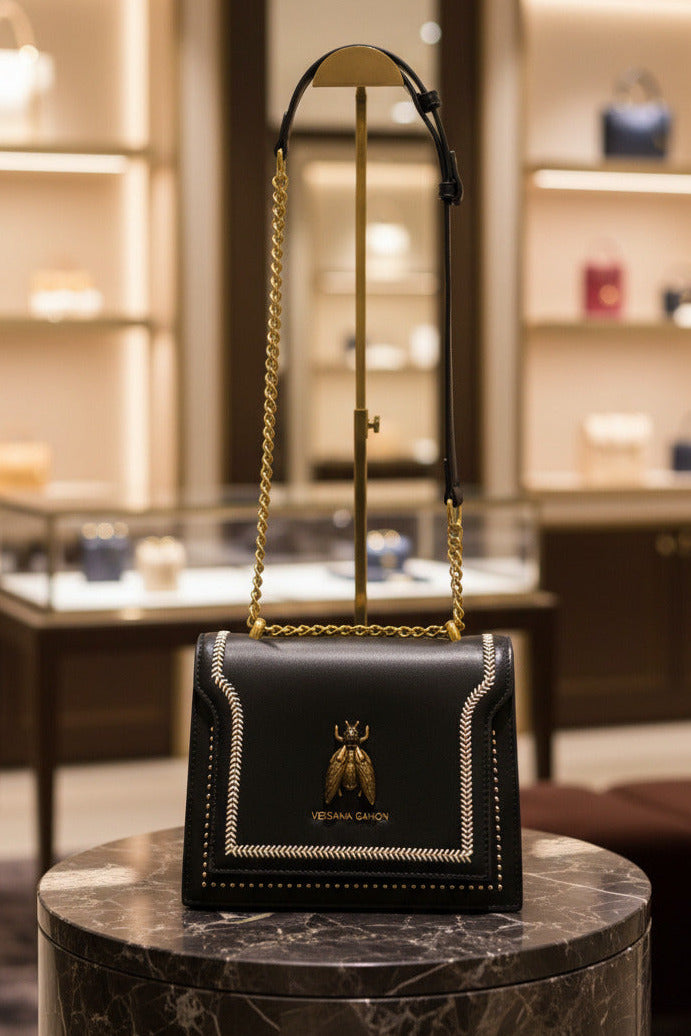 A black Vessana Gahon crossbody bag with a gold chain strap, an adjustable black leather shoulder strap, and a gold bee logo clasp on a gray carpeted floor.