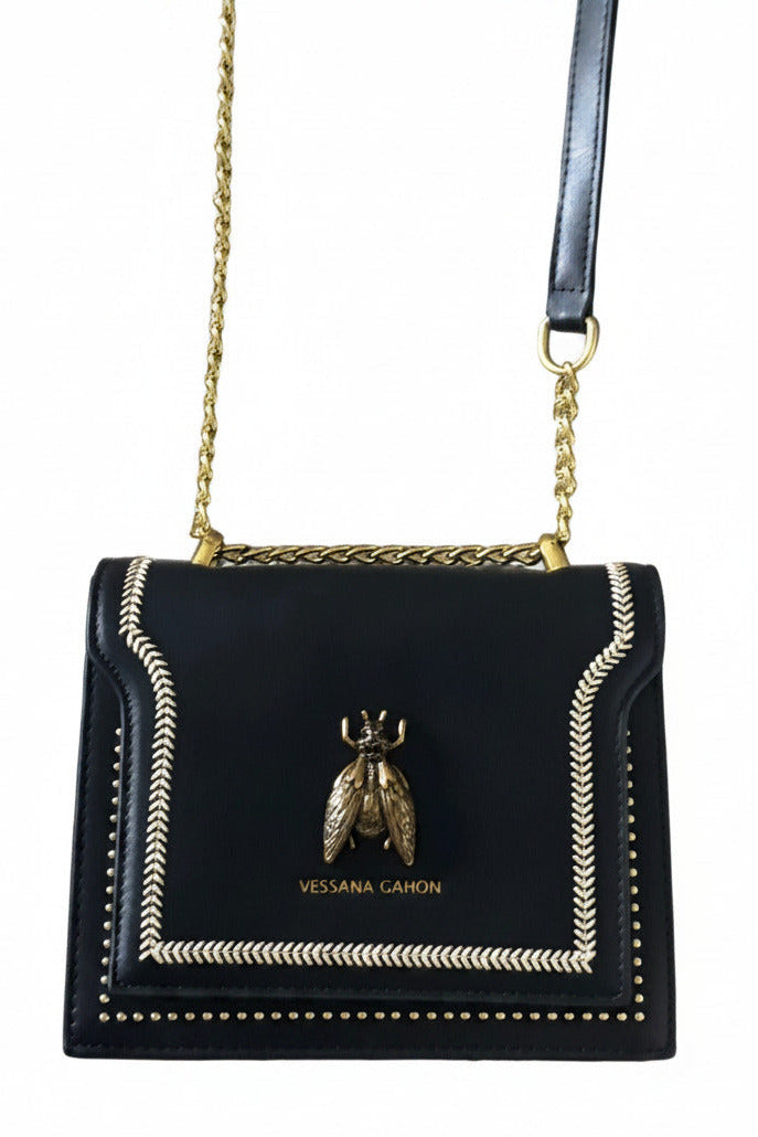 A black Vessana Gahon crossbody bag with a gold chain strap, an adjustable black leather shoulder strap, and a gold bee logo clasp on a gray carpeted floor.