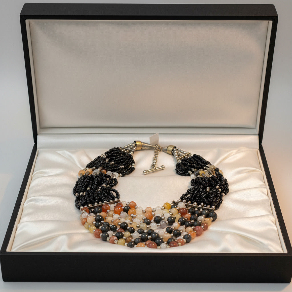 A multi-strand beaded necklace featuring black seed bead strands, silver-toned spacer beads, and a tiered bottom section with polished beads in black, gray, white, orange, and yellow, shown on a plain white background.