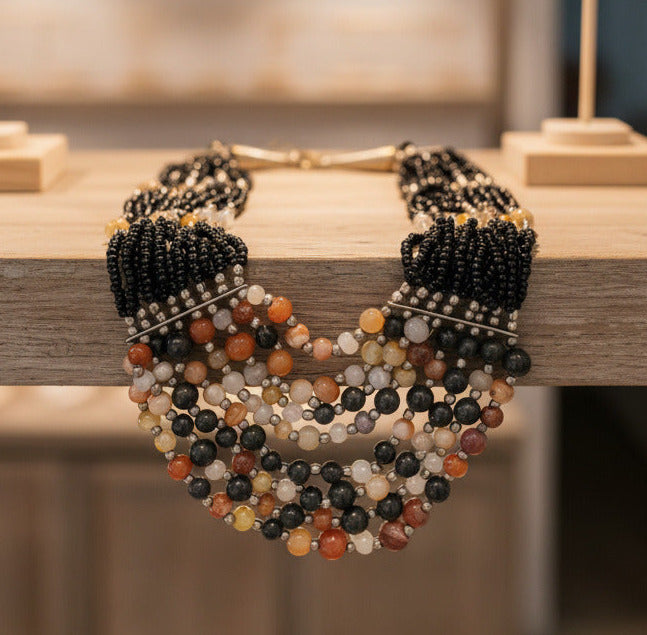 A multi-strand beaded necklace featuring black seed bead strands, silver-toned spacer beads, and a tiered bottom section with polished beads in black, gray, white, orange, and yellow, shown on a plain white background.