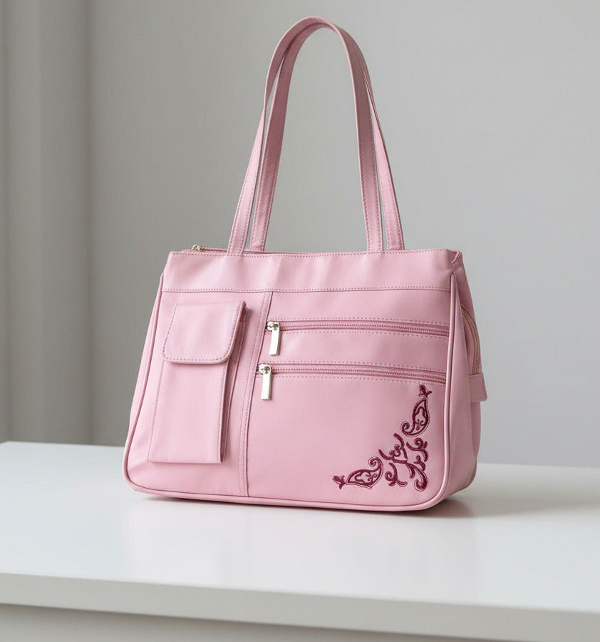 A pink nylon shoulder bag with a long strap, featuring a front pocket, two zippered compartments, and dark pink embroidered scrollwork in the bottom right corner.