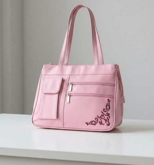 A pink nylon shoulder bag with a long strap, featuring a front pocket, two zippered compartments, and dark pink embroidered scrollwork in the bottom right corner.