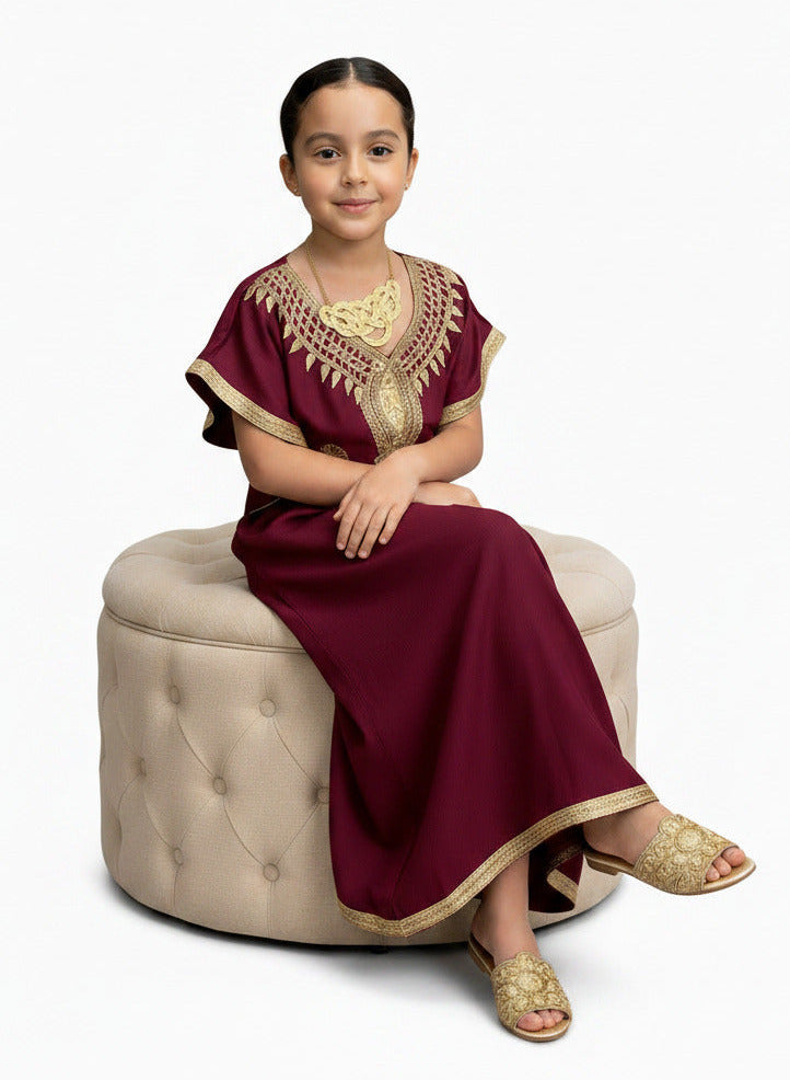 A young girl modeling a long maroon kaftan dress with gold embroidery on the neckline and hem, standing against a plain white background.