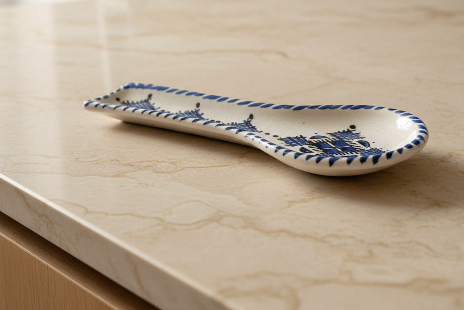 A ceramic spoon rest with a cream-colored glaze, decorated with dark blue geometric patterns and a braided border, shown on a white kitchen counter. 