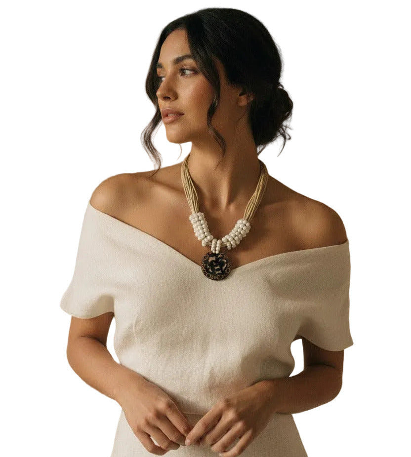 A bohemian-style necklace featuring a large round black pendant with white mother-of-pearl floral cutouts and brown accents, suspended from multiple beige cords with white beads.