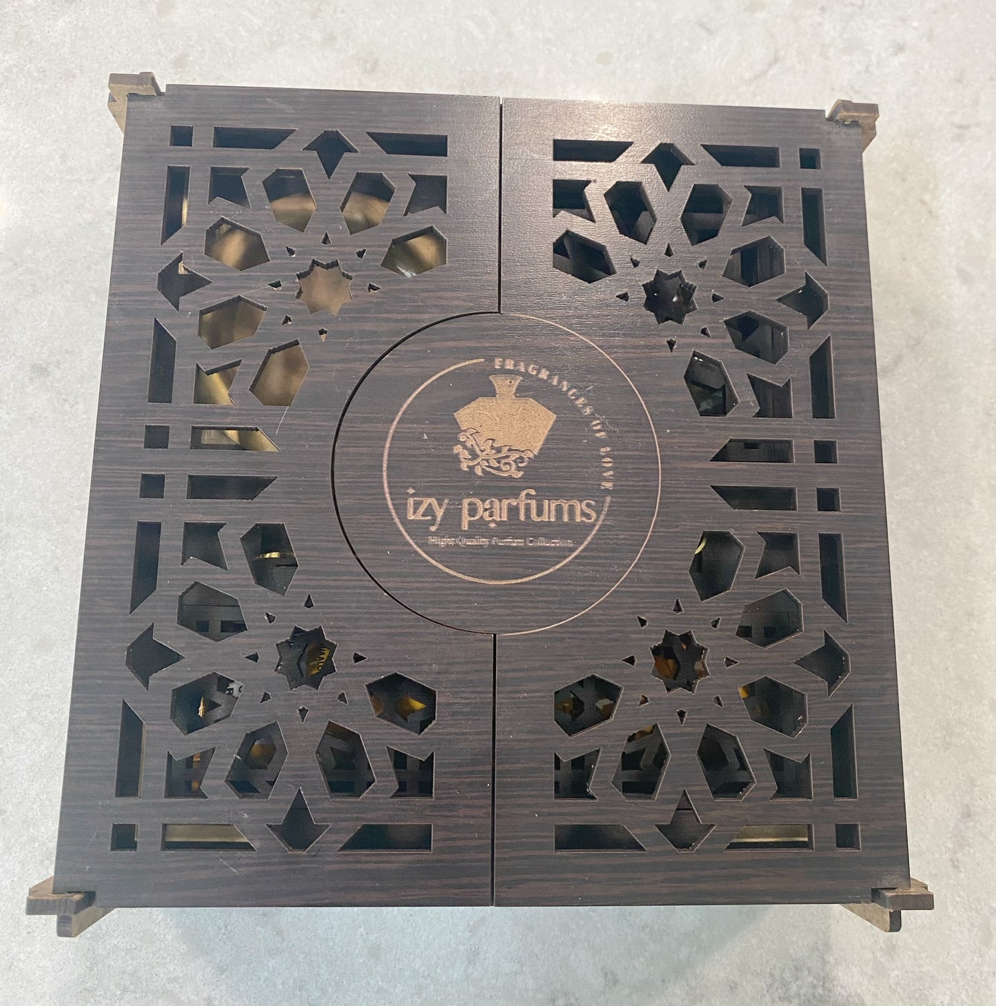 A square dark brown wooden box with intricate geometric cutout patterns on the lid, featuring a central circular logo for 'Izy parfums' --TTP