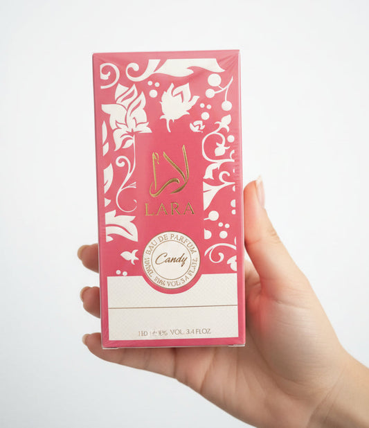 A hand holding a pink rectangular perfume box with white floral cutout-style patterns and the brand name LARA visible on the front, photographed on a white surface.